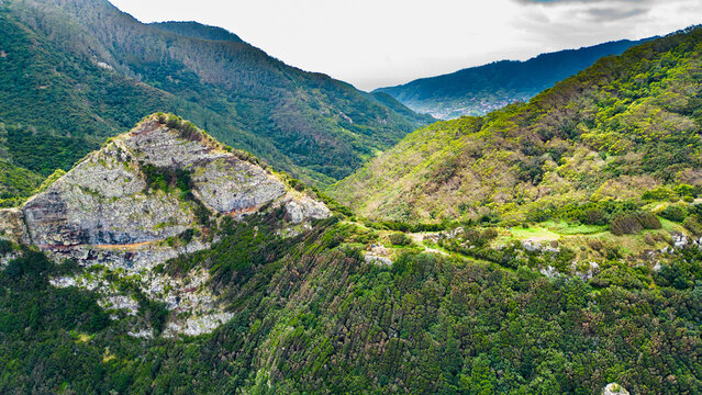 Aerial view of Madeira's lush mountains and valleys. Green slopes, a distinctive rocky peak, and distant village hint at island's natural beauty and hiking trails. Vereda do Larano