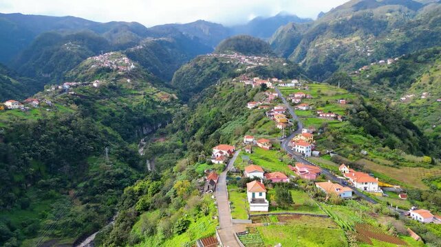 Aerial view of a picturesque village Faial nestled in Madeira's verdant mountains, with houses clinging to terraced slopes under a cloudy sky.