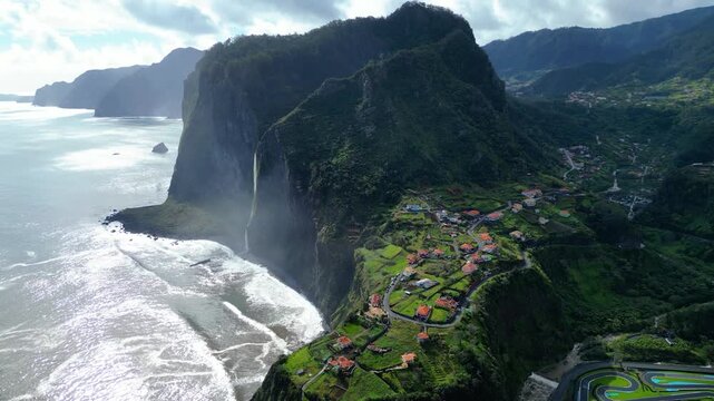 Aerial view of Madeira's rugged coastline near Faial, featuring a dramatic waterfall plunging into the Atlantic, a charming village  and terraced landscapes under a dynamic sky.