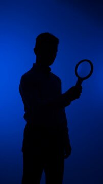 Vertical video man silhouette performing an investigation using a magnifying glass, depicting concepts of discovery, search, and research against a dark blue background