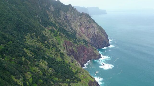 Dramatic aerial view of Madeira's lush green cliffs plunging into the turquoise Atlantic, with waves crashing against the rugged coastline under an overcast sky. Vereda do Larano