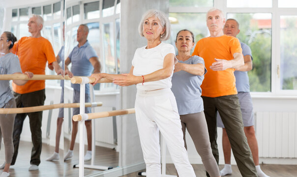 Positive old female attendee of ballet courses doing battement tendu standing by training barre with others