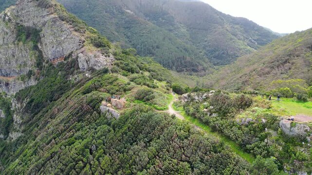 Aerial view of Madeira's lush mountains and valleys. Green slopes, a distinctive rocky peak, and distant village hint at island's natural beauty and hiking trails. Vereda do Larano