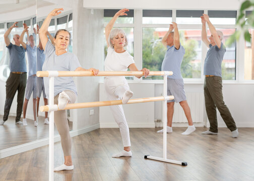 Elderly women with group of dancers practicing ballet pose grand batman near ballet barre in modern dancing hall