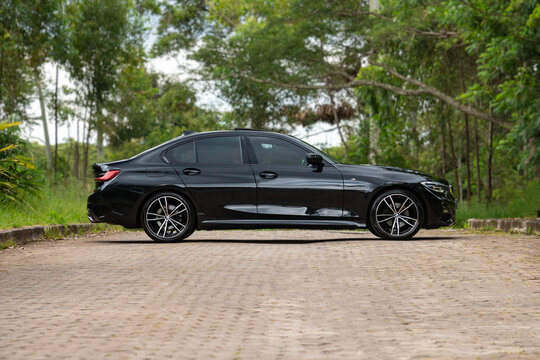 Black BMW 320i full side profile view in front of forest, luxury family car on paved road near trees