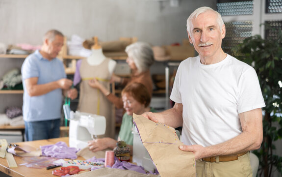 Elderly male participant present fabric billet, study sewing in educational coterie. Some people communicate and discuss work, while others sew using sewing machine, perform task with mannequin. .