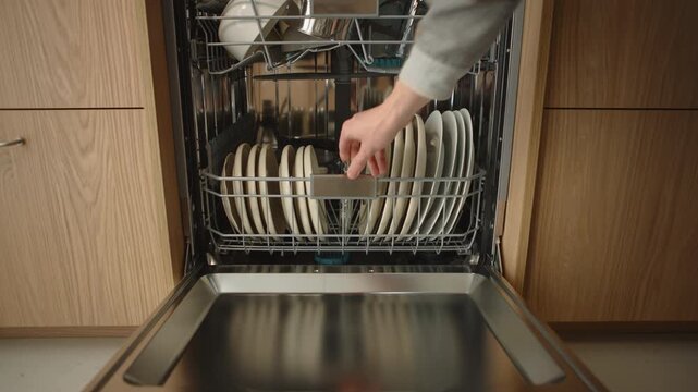 Male hand closes built-in dishwasher door in modern kitchen. Wooden cabinets, organized tableware and home interior details highlight everyday household routine and appliance use in residential space