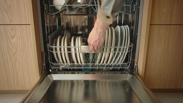 Caucasian man closes dishwasher door in modern kitchen. Wooden cabinets, organized tableware and home interior details highlight everyday household routine in residential space. Daylight, close-up
