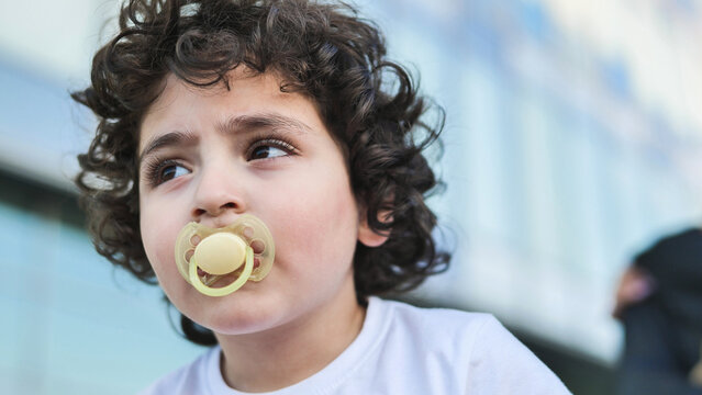 Little child with curly hair and pacifier innocently looking away outdoors