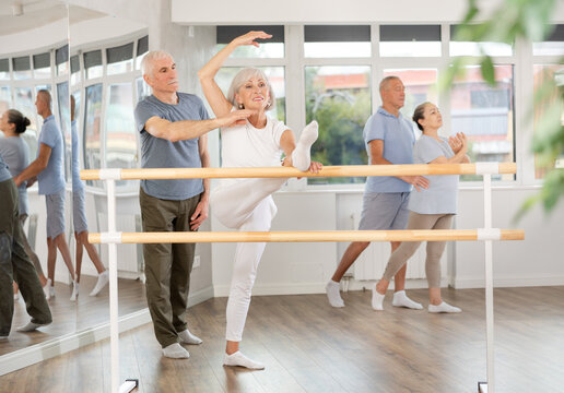 Positive old female attendee of ballet courses stretching her leg standing by training barre under control of instructor