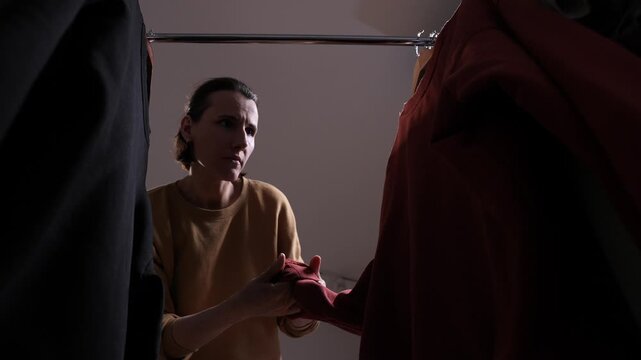 Woman standing in front of clothing rack, holding a garment, feeling confused and worried while making a difficult decision about what to wear in a challenging situation