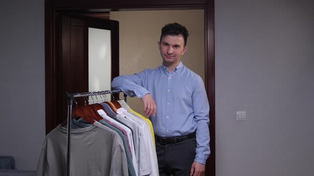 Man leaning on a clothing rack of colorful t shirts and casual shirts, smiling confidently while choosing outfit options in a modern indoor boutique or studio setting