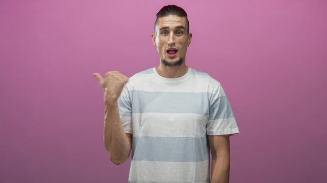 Man pointing thumb over shoulder with open mouth and visible beard in studio wearing striped tee, facing camera; surprise curiosity invitation.