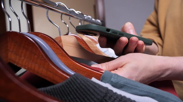 Person holding a smartphone, scanning a barcode tag on a garment hanging in a closet or retail rack, engaging in modern shopping or inventory management