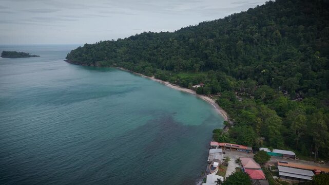Aerial drone view of tropical beach, sea and Thai houses surrounded by lush forest. Scenic coastal landscape in Thailand with turquoise water and traditional buildings from above.