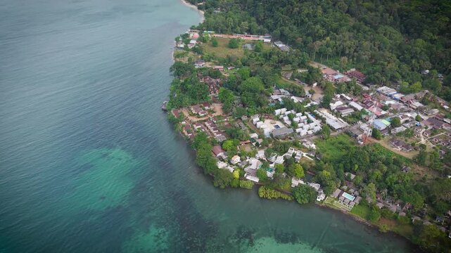 Aerial drone view of tropical beach, sea and Thai houses surrounded by lush forest. Scenic coastal landscape in Thailand with turquoise water and traditional buildings from above.