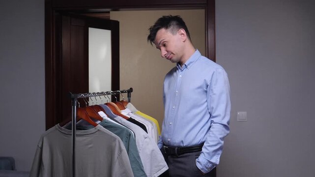 Man feeling confused and indecisive while looking at a clothing rack with various t shirts, gesturing with his hands in a shrug, struggling with outfit choice