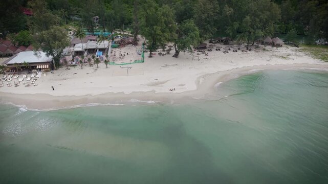 Aerial drone view of tropical beach, sea and Thai houses surrounded by lush forest. Scenic coastal landscape in Thailand with turquoise water and traditional buildings from above.