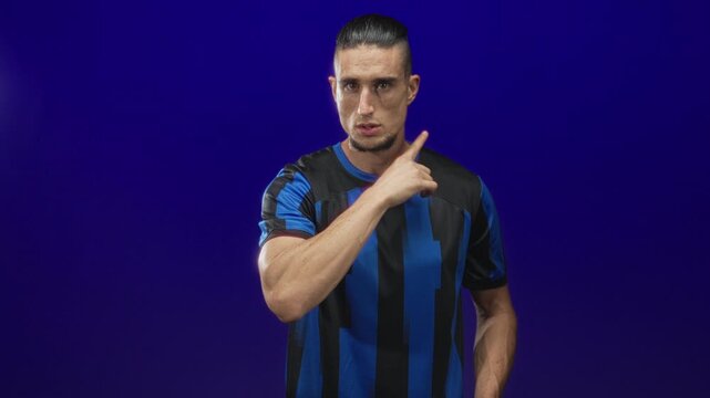 Man in blue and black soccer jersey points finger at camera with stern gaze and assertive pose against a plain studio backdrop; determination challenge focus.