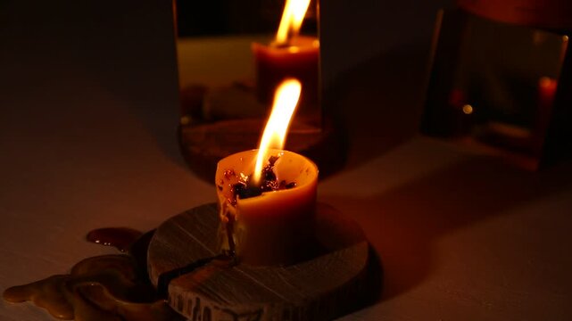 A brightly wind burning candle against a dark background near a mirror, the candle flame dripping melted wax onto the table. A magical witch ritual.