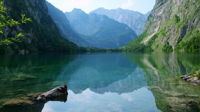 The video shows Obersee, a mountain lake in the Alps, Bavaria, Germany. The water reflects the surrounding mountains and trees. Visitors enjoy the natural beauty and landscape.