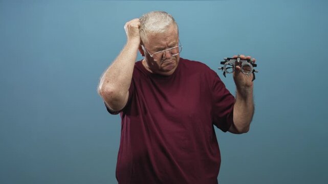 Man holding trial frame and scratching head in studio while wearing eyeglasses and a maroon shirt; confusion aging.