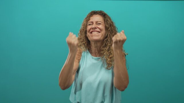 Middle aged hispanic woman clapping hands and pumping fists with a big smile against a turquoise studio backdrop; joy celebration.