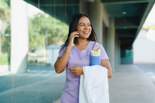 Young hispanic female healthcare professional wearing scrubs, talking on a smartphone and laughing while walking outside a medical building
