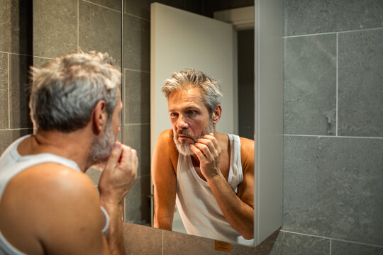 Middle-aged man inspecting beard in home bathroom mirror