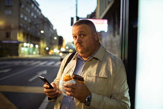 Middle-aged man with coffee and phone at city bus stop at dawn