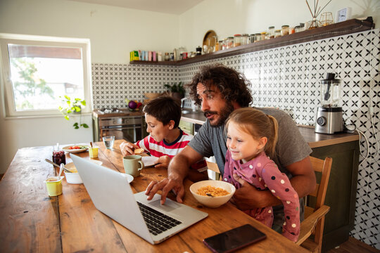 Father working on laptop with children during breakfast in home kitchen