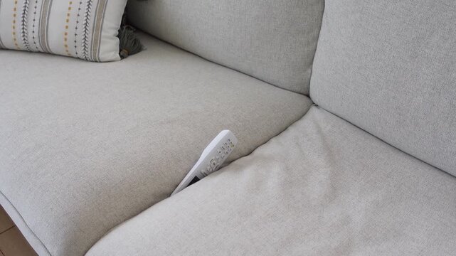White air conditioning remote control tucked into corner of gray sofa in bright living room.