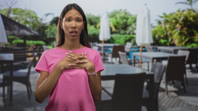 Thai woman clasping hands over chest with closed eyes in outdoor poolside cafe by swimming pool at building; relief reflection.