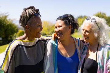 Fototapeta premium Diverse female friends including senior laughing and hugging in park with sunglasses and shawls