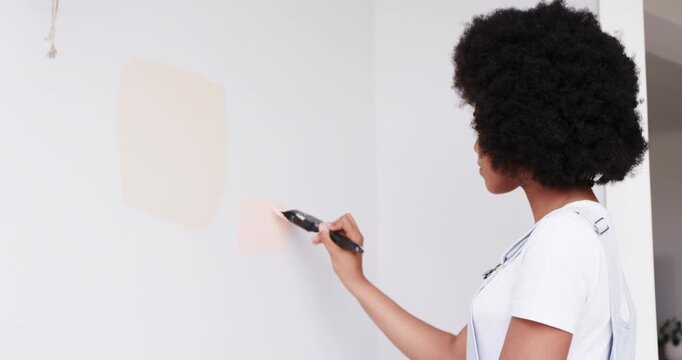 African American woman in overalls applying peach swatch with brush in room, evaluating, copy space