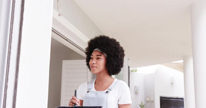 African American woman on porch in overalls surveying above door touching up frame with brush, tray