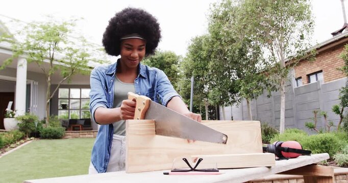 Woman placing board in miter box, sawing with handsaw in backyard wearing denim shirt for DIY