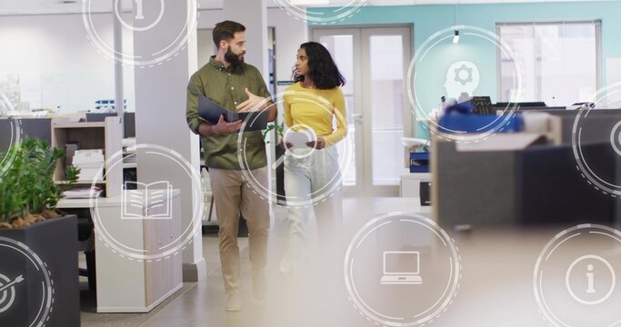 Walking coworkers wearing mustard top, gesturing through office aisle, holding folio and tablet