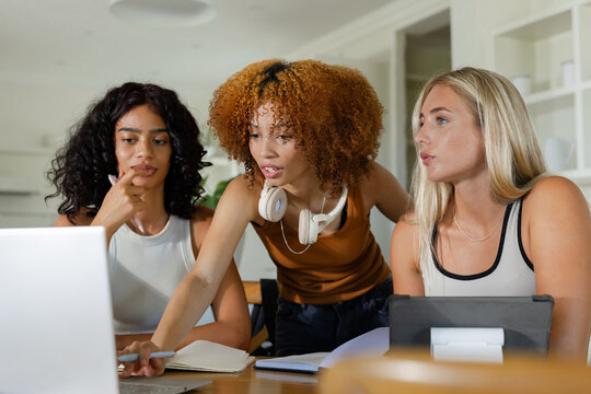 Three female friends collaborating around table in kitchen, pointing at laptop wearing headphones