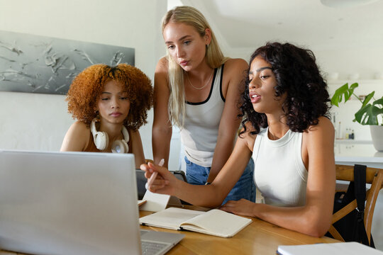 Diverse female friends collaborating around laptop at home kitchen table with notebook, headphones