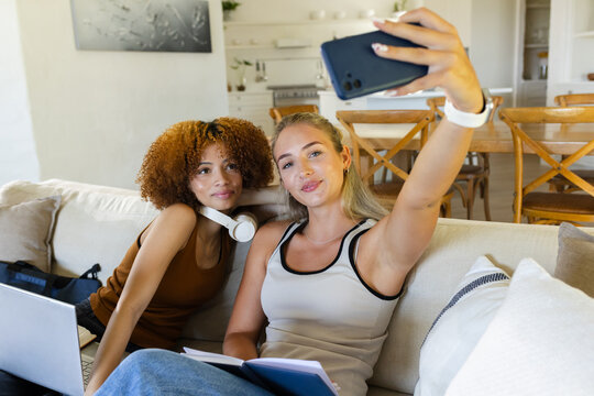 Two female friends posing and taking smartphone selfie on sofa in bright open-plan living area