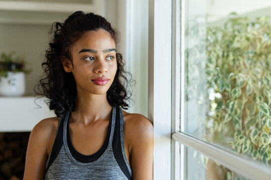 Adult African woman standing by home window, gazing outside near plant and firewood, in grey tank