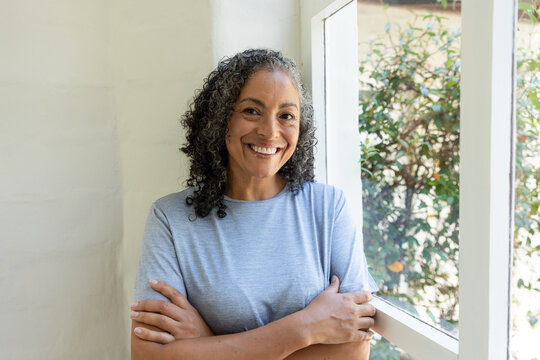 African American woman standing at home by window, leaning against white frame, wearing gray tee