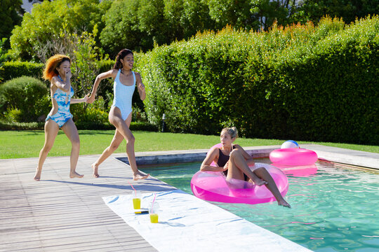 Diverse female friends in swimsuits running hand-in-hand at backyard pool with pink rings, towel