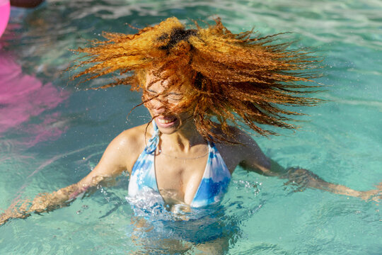 Adult woman flipping hair in turquoise pool in blue halter bikini top, droplets flying past float