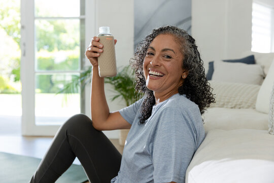 African American woman sitting on floor in living room holding beige bottle in workout top smiling