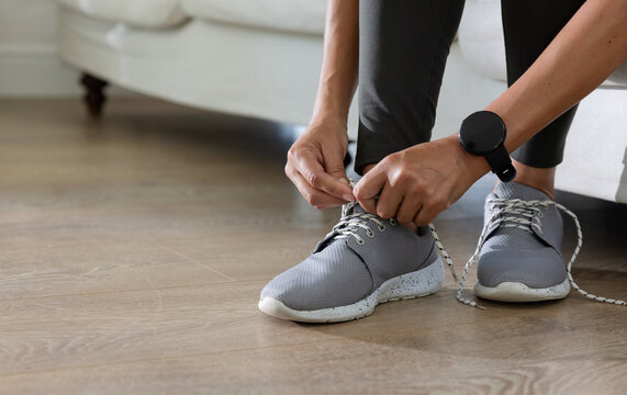 African American woman on sofa tying gray sneakers with laces, wearing dark leggings and smartwatch