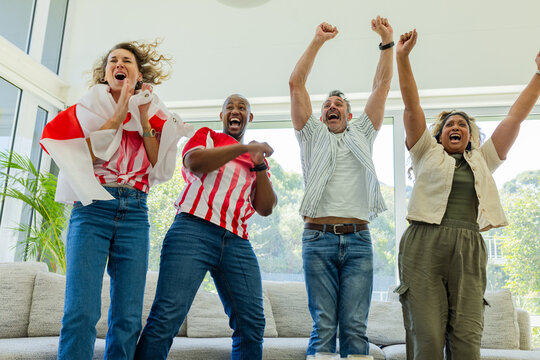 Diverse friends standing on couch at home, cheering, waving red white flag, wearing jersey