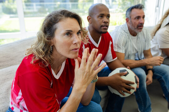 Diverse friends wearing red jerseys watching soccer on sofa in bright living room with soccer ball