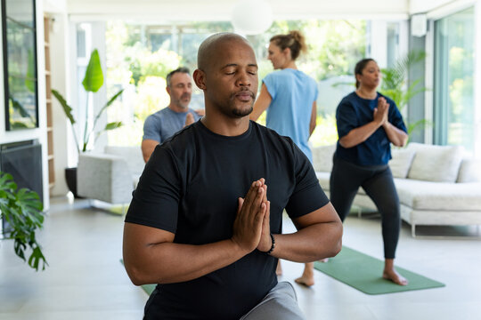 Diverse group wearing activewear practicing yoga and meditating in living room on mats by sofa